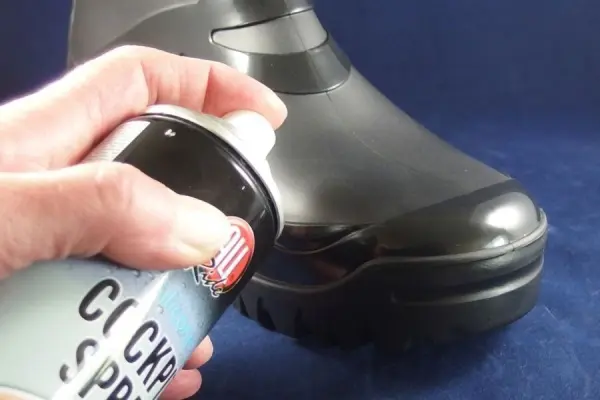 Ein schwarzer Gummistiefel wird mit Cockpitspray poliert, während eine Hand die Spraydose hält. Der Hintergrund ist dunkel.