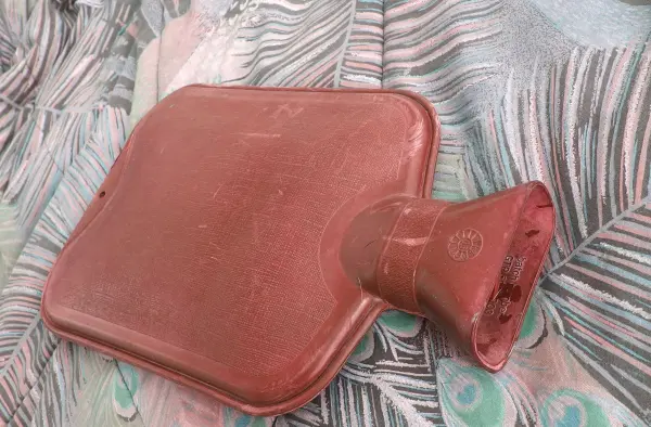 Eine rote Wärmflasche liegt auf einem bunten, gemusterten Untergrund und ist bereit, länger warmes Salz zu halten.