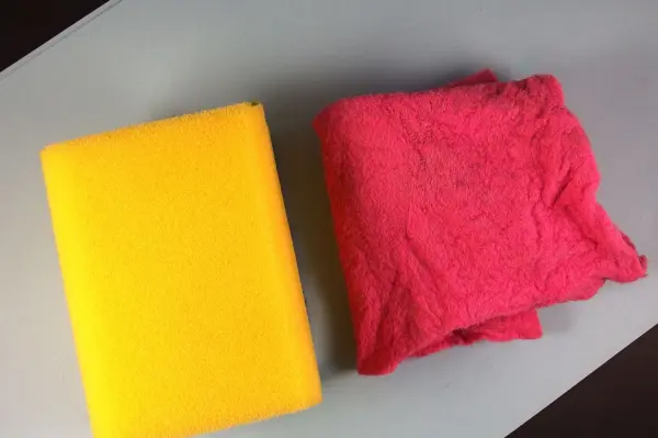 Ein gelber Schwamm und ein rosa Spüllappen liegen auf einer grauen Fläche, bereit zur Reinigung in der Mikrowelle.