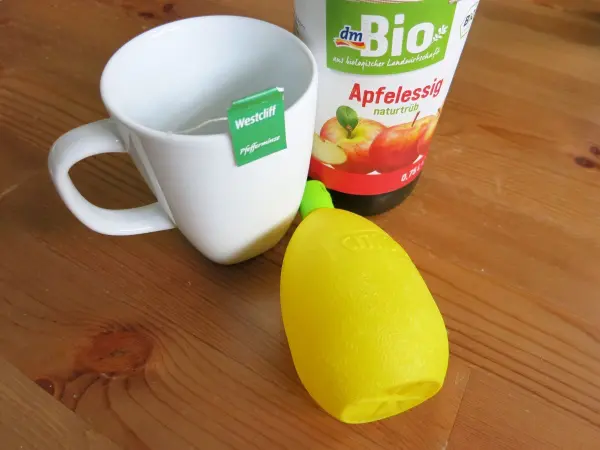 Eine Tasse mit Teebeutel steht neben einer Flasche Apfelessig und einer gelben Zitronenpressform auf einem Holzuntergrund.