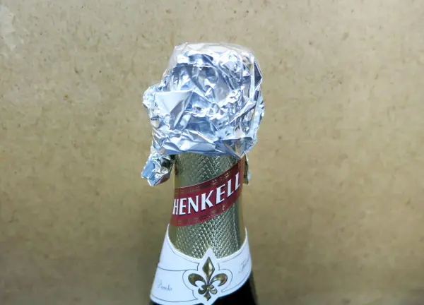 Eine Sektflasche mit Aluminiumfolie als Verschluss bedeckt, steht vor einem neutralen Hintergrund. Text: „HENKELL“.