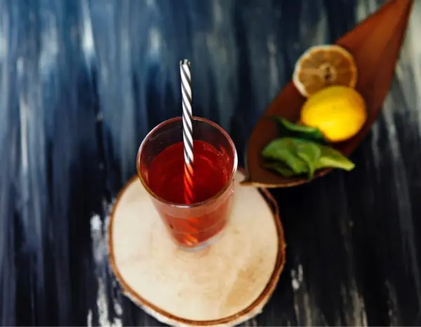 Ein Glas mit rotem Getränk und gestreiftem Strohhalm steht auf einem Holzbrett. Daneben liegen Zitrone und Limettenblätter.