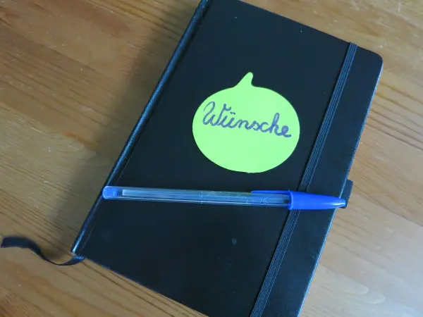 Ein schwarzes Notizbuch mit einem grünen Haftnotizzettel und einem blauen Stift liegt auf einem Holztisch.