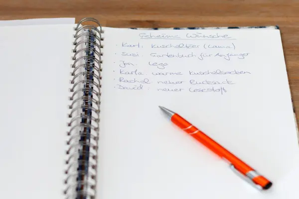 Ein Notizbuch liegt offen auf einem Tisch mit der Überschrift "Geheime Wünsche", begleitet von einem orangefarbenen Stift.