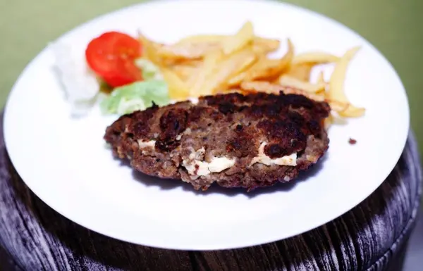 Ein gegrilltes Bifteki liegt auf einem weißen Teller, begleitet von Pommes und einem frischen Salat mit Tomate.