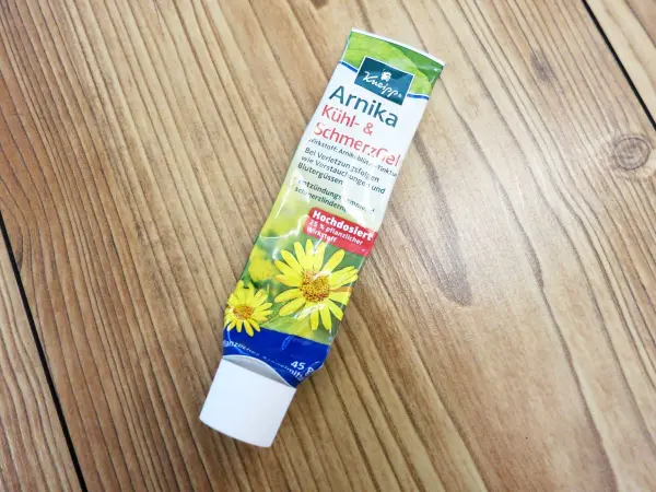Eine Tube Arnikasalbe steht auf einem Holzfußboden, bereit zur Anwendung gegen Mückenstiche. Die Verpackung zeigt gelbe Blumen.