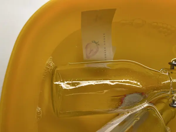 Eine Flasche liegt in einer gelben Schüssel mit Orangenreiniger, während ein Etikett im Wasser schwebt und sich löst.