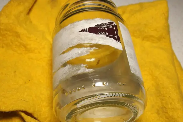 Ein Glas mit Kleberesten liegt auf einem gelben Tuch, während ein Orangenreiniger verwendet wird, um die Etiketten zu entfernen.