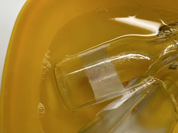 Eine Glasflasche liegt flach in einem gelblichen Orangenreiniger, der Klebereste aufweicht.
