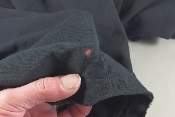 Ein Hand hält ein schwarzes Kleidungsstück mit einem kleinen Loch, was auf Mottenschäden hinweist. Ein Hinweis auf „Motten“.