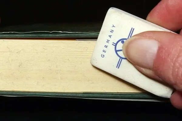 Ein Buch liegt auf einem Tisch, während eine Hand einen Radiergummi benutzt, um Schmutz vom Buch zu beseitigen.