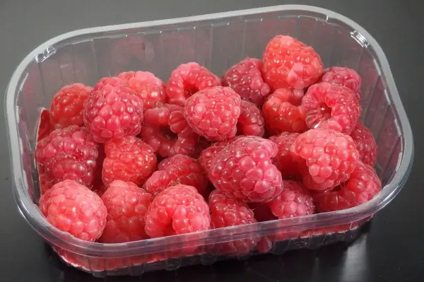Frische Himbeeren liegen in einer transparenten Plastikschale auf einer dunklen Oberfläche, bereit zum Verzehr.