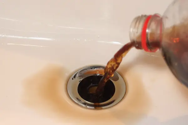 Eine Flasche Cola wird in ein Abflussloch gegossen, während die braune Flüssigkeit durch den Abfluss fließt.