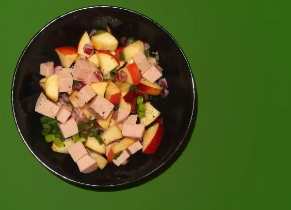 Wurstsalat mit Apfelstücken und Zwiebeln in einer schwarzen Schüssel auf grünem Hintergrund.