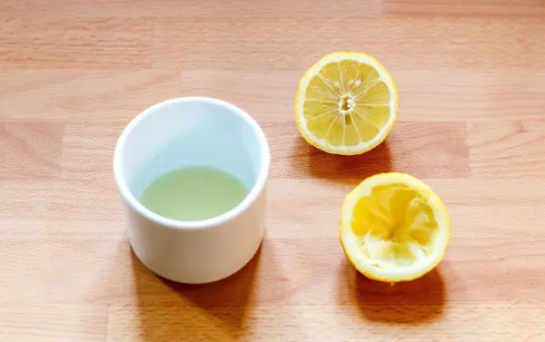Eine halbe Zitrone und ein Becher mit Zitronensaft stehen auf einem Holztisch, bereit zur Anwendung gegen Obstfliegen.