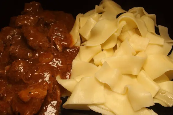 Ein reichhaltiges Gulasch liegt neben frisch zubereiteten Bandnudeln auf einem schwarzen Teller, appetitlich angerichtet.