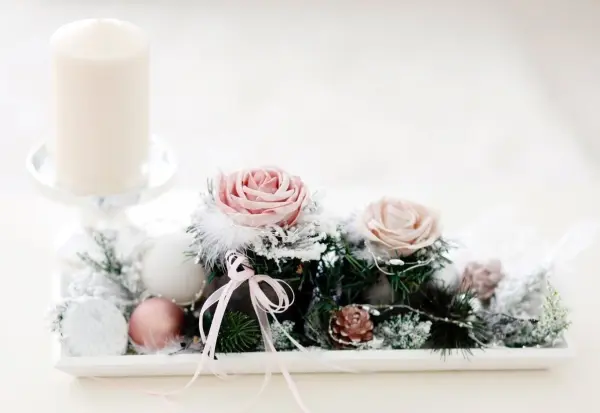 Ein dekorativer Tischschmuck mit einer weißen Kerze, zarten Rosen und Tannenzweigen, ideal zum Frischhalten einer Tanne.
