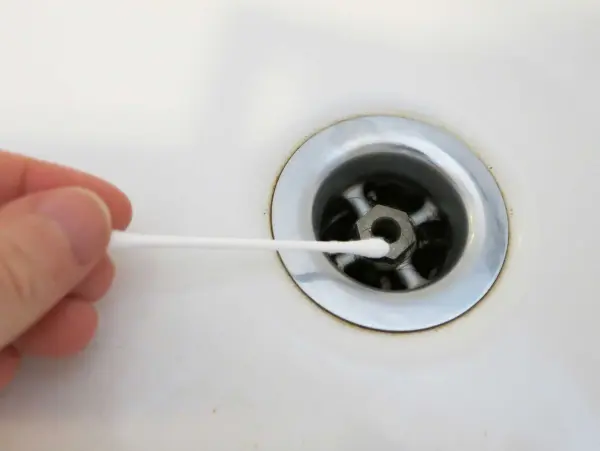Ein Wattestäbchen wird in den Abfluss einer Badewanne gehalten, um Haare zu entfernen. Der Hintergrund ist eine weiße Badewannenoberfläche.