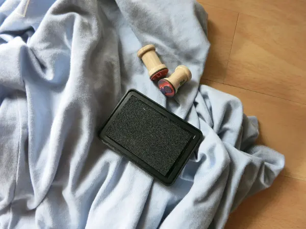 Stempelkissen liegt auf einem blauen Stoff, daneben zwei Holzstempel, auf hellem Holzboden platziert.
