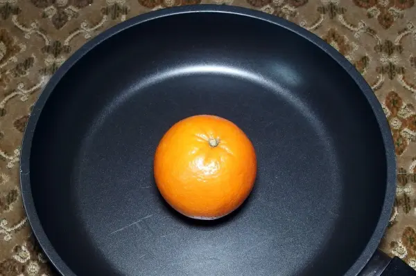 Eine orangefarbene Orange liegt in einer schwarzen Pfanne auf einem gemusterten Untergrund.