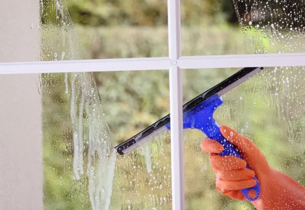 Hand reinigt Fenster mit blauem Abzieher, Seifenreste auf Glas, grüne Natur draußen.