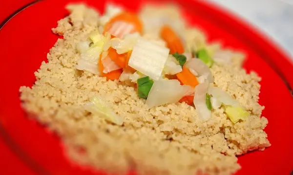 Leckerer Gemüse-Couscous liegt auf einem roten Teller. Bunte, geschnittene Gemüsewürfel sind darüber verteilt.