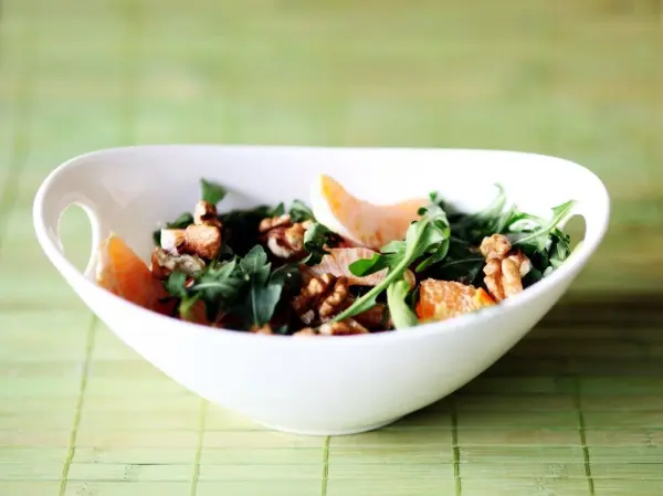 Ein frischer Ruccola-Mandarinen-Salat mit Walnüssen liegt in einer eleganten weißen Schüssel auf einem grünen Untergrund.