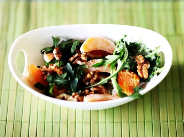 Ein Salat aus Rucola und Mandarinen wird mit Walnüssen in einer weißen Schüssel angerichtet und auf einem grünen Untergrund präsentiert.