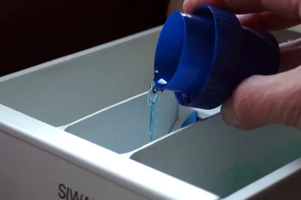 Eine Hand gießt flüssiges Waschmittel aus einem blauen Behälter in ein Waschmittelfach zum Vermeiden von muffiger Wäsche.