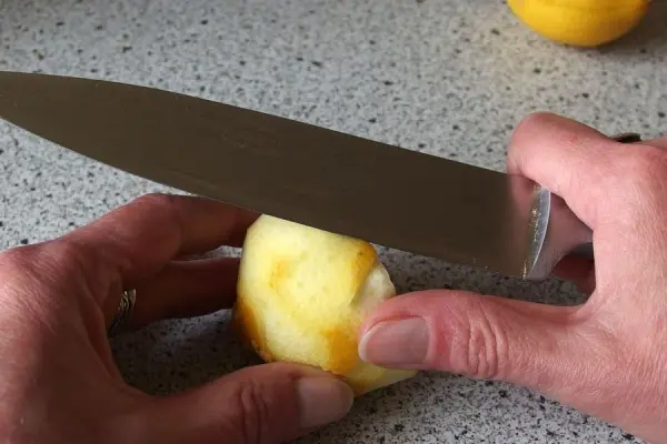 Ein Messer schneidet die Zitronenschale, während die Hände die Zitrone halten. Eine helle Arbeitsfläche umgibt die Szene.