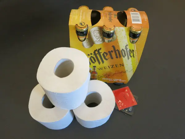 Drei Toilettenrollen liegen auf einem Tisch, während im Hintergrund eine Kiste mit Getränken steht.