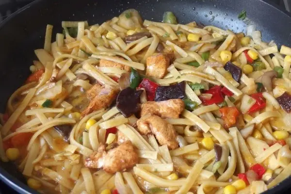 Gebratene Nudeln werden in einer Pfanne zubereitet, vermischt mit buntem Gemüse und Fleischstücken, aromatisch und appetitlich.