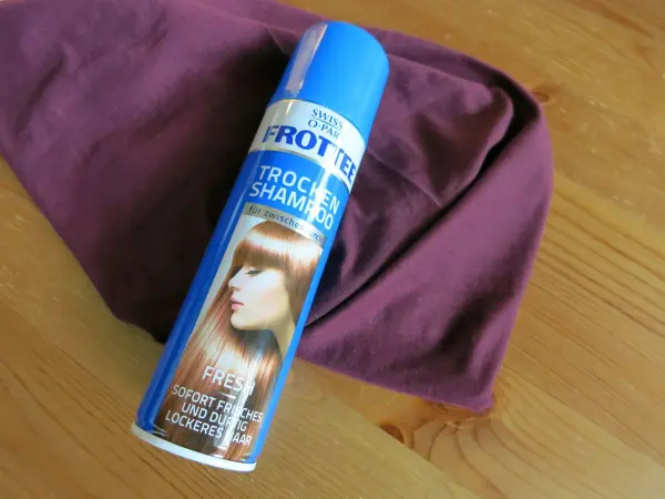Eine Flasche Trockenshampoo liegt auf einem lila Kleidungsstück auf einem Holztisch, um Fettflecken zu entfernen.