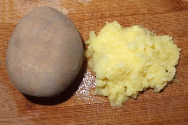 Eine rohe Kartoffel liegt neben geriebenen Kartoffeln auf einem Holzbrett, bereit für die Zubereitung von Klößen.