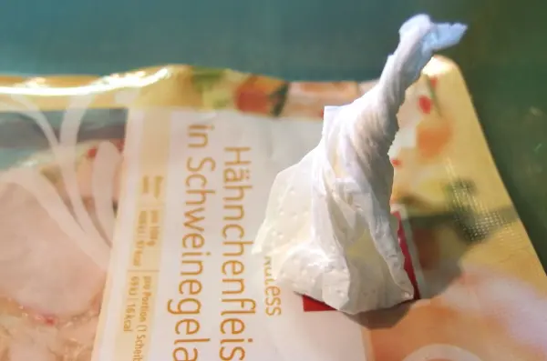 Eine kleine, feuchte Papierserviette steht auf einer Verpackung für Aufschnitt. Sweat in Kühlschrank-Aufschnittdosen vermeiden wird thematisiert.