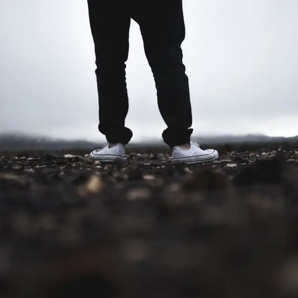 Weiße Stoffturnschuhe stehen auf einem schmutzigen Untergrund, während eine graue, wolkige Landschaft im Hintergrund sichtbar ist.