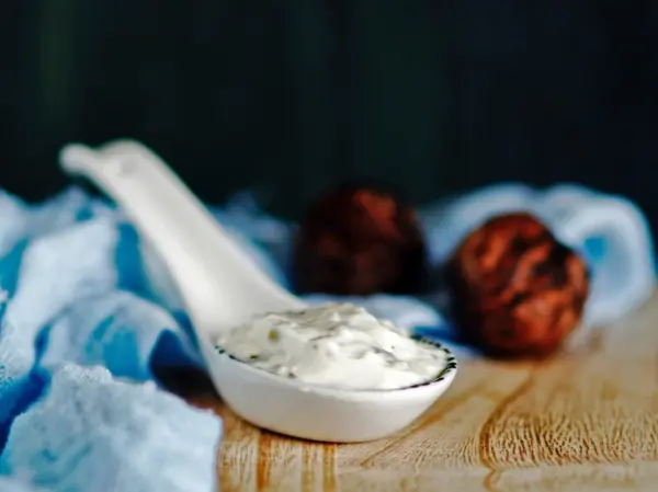 Ein cremiger Edel-Feta-Dip liegt in einem Löffel, umgeben von blauen Stoff und zwei dunklen, runden Snacks.