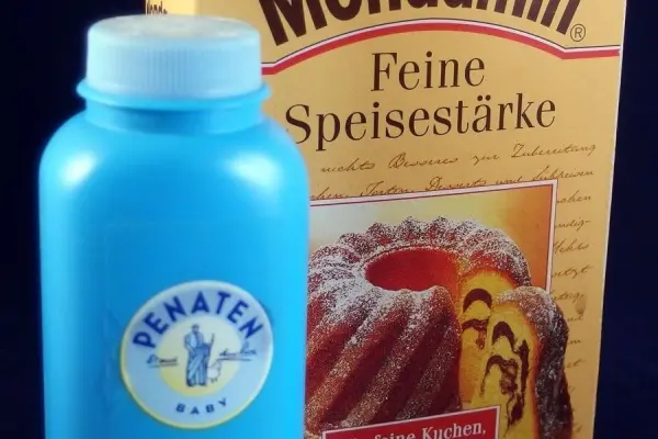 Eine blaue Flasche mit Babypuder steht neben einer Packung Speisestärke, die auf einem dunklen Untergrund platziert ist.