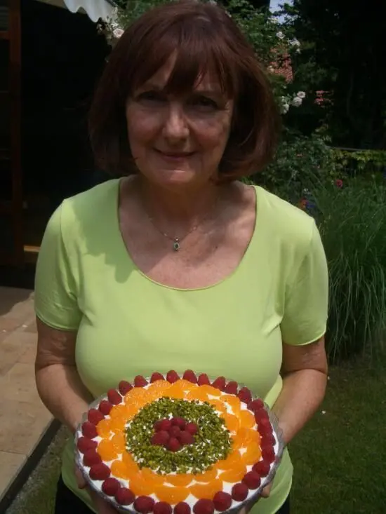 Eine Frau hält stolz einen bunten Sherry Himbeer Trifle in einem Garten, umgeben von grünen Pflanzen und Sonne.