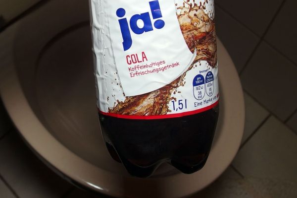Eine Cola-Flasche schwebt über einer Toilette, bereit zum Reinigen der WC-Schüssel mit dem Getränk.