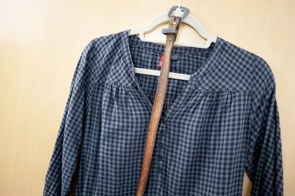 Eine lange Bluse hängt an einem Kleiderbügel neben einem Stock an einer hellen Wand, bereit zum Tragen oder Stylen.