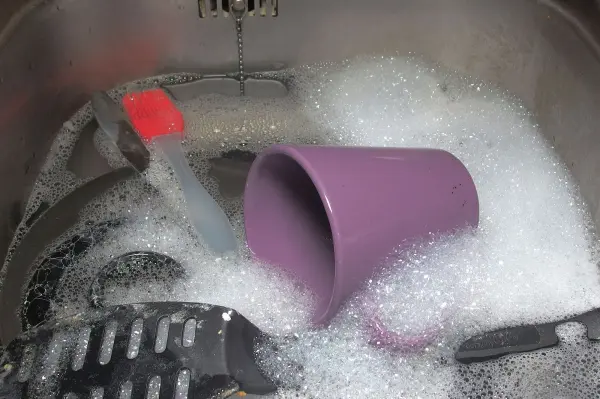 Ein lila Becher liegt schräg in einer Spülmaschine, umgeben von Schaum und Geschirr, während die Klappe der Maschine offen ist.