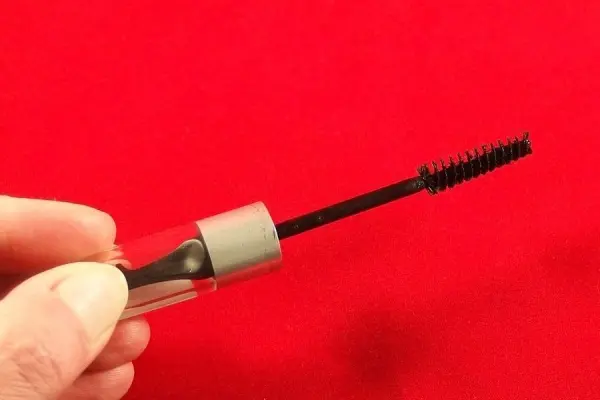 Eine Hand hält eine Wimperntusche hoch, während sie vor einem roten Hintergrund gezeigt wird, was auf eingetrocknete Wimperntusche hinweist.