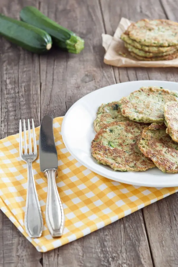 Zucchinipuffer auf weißem Teller liegen auf rustikalem Holztisch neben einem gelben Tischset, Zucchini im Hintergrund.