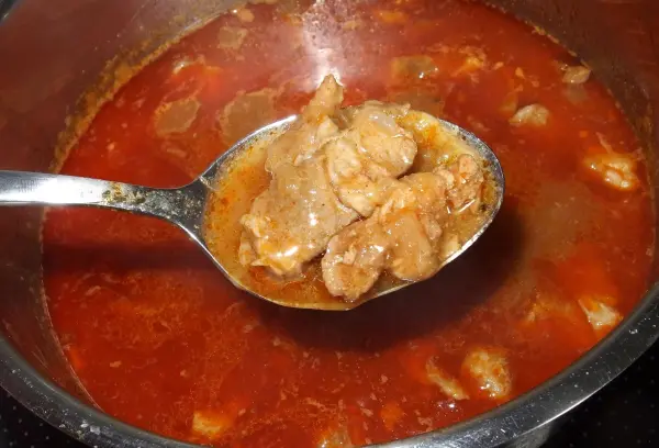 Ein Löffel mit zartem Fleisch in einer dampfenden, roten Soße wird aus einem großen Topf entnommen, der auf einem Herd steht.
