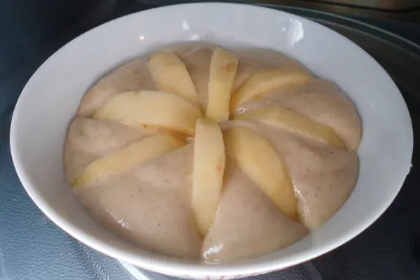 Ein ungebackener Apfelkuchen mit Apfelscheiben sitzt in einer weißen Schüssel, umgeben von einer cremefarbenen Teigmasse.