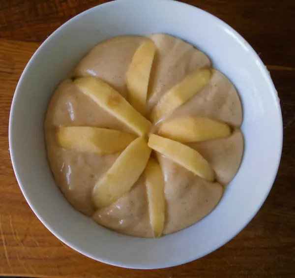 In einer weißen Schüssel liegt Apfelkuchen-Teig, darauf sechs Apfelschnitze strahlenförmig angeordnet auf Holztisch.