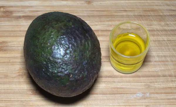 Eine Avocado liegt neben einem kleinen Glas mit Olivenöl auf einer Holzoberfläche, bereit zur Haarpflegeanwendung.