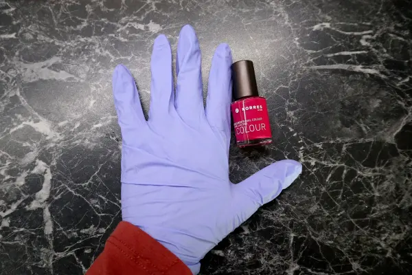 Eine Hand mit blauen Handschuhen liegt auf einem Tisch, während daneben eine Flasche Nagellack in leuchtendem Pink steht.