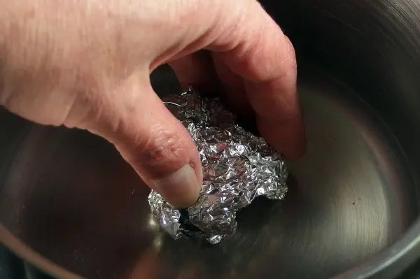 Eine Hand greift in einen Topf mit Wasser, während sie ein Stück Alufolie zum Scheuern verwendet.